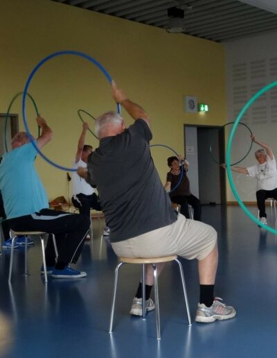 Ältere Menschen sitzen in einem Kreis auf einem Hocker. Sie halten einen Hoola-Hoop-Reifen in den Händen und lehnen sich nach links.